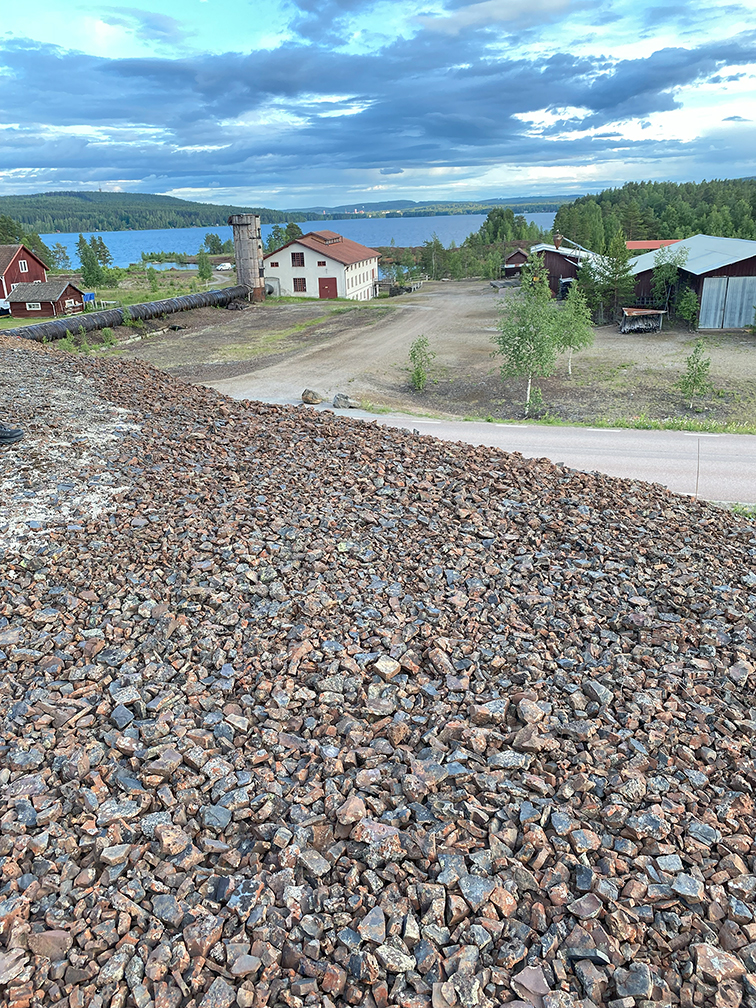 View from a slag heap towards houses with a lake in the background and forested hills at the horizon under ragged clouds